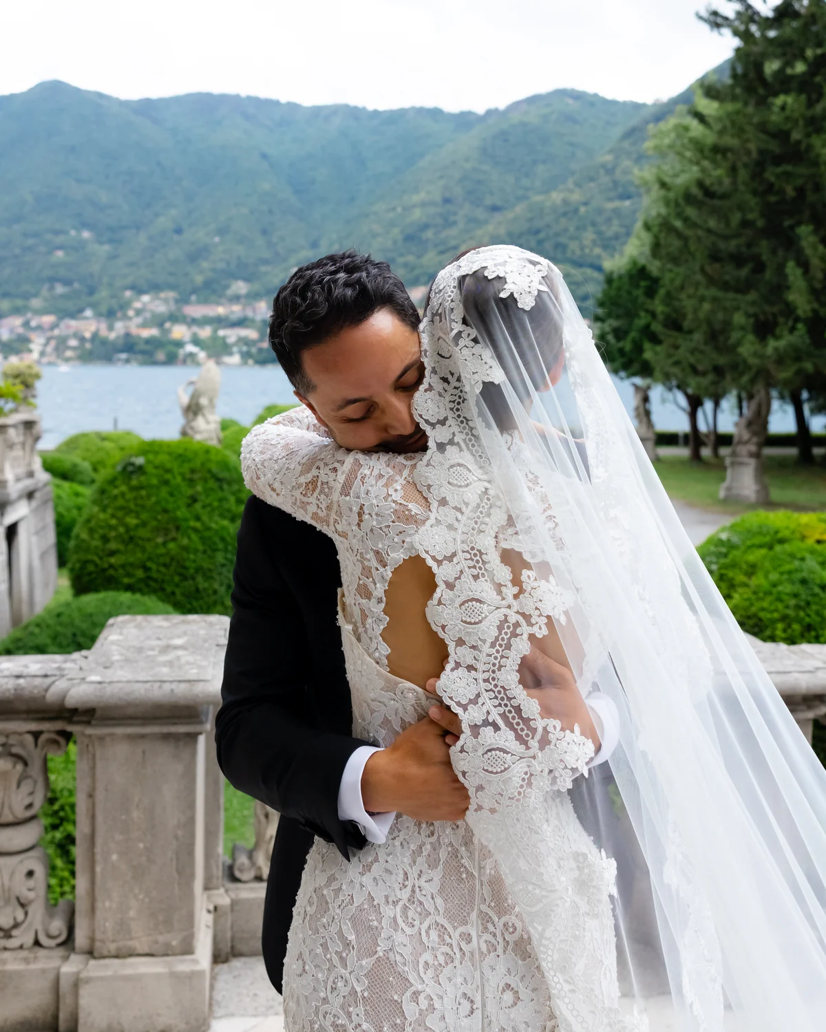 Groom embracing the bride in her long-sleeve lace gown and veil — Lee Petra Grebenau Versailles at Lake Como