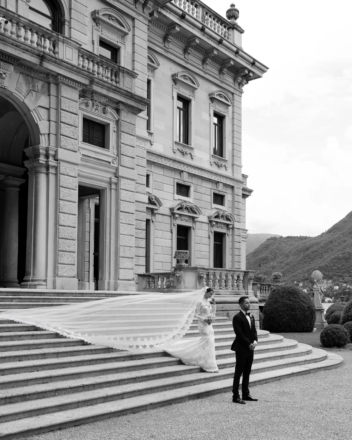 Bride's cathedral train cascading down the Villa Erba staircase, groom below — Lee Petra Grebenau Versailles gown